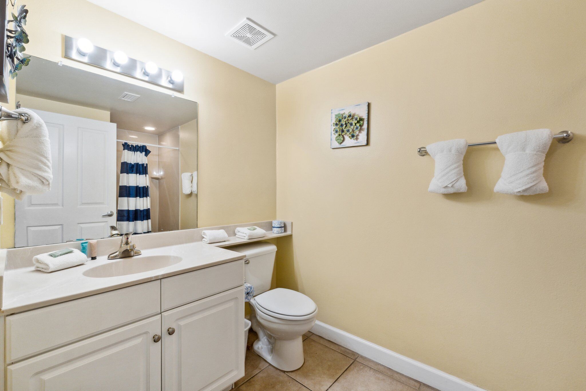 A bathroom with a sink, toilet, and a mirror with lights. There are towels on the wall and a shower curtain.