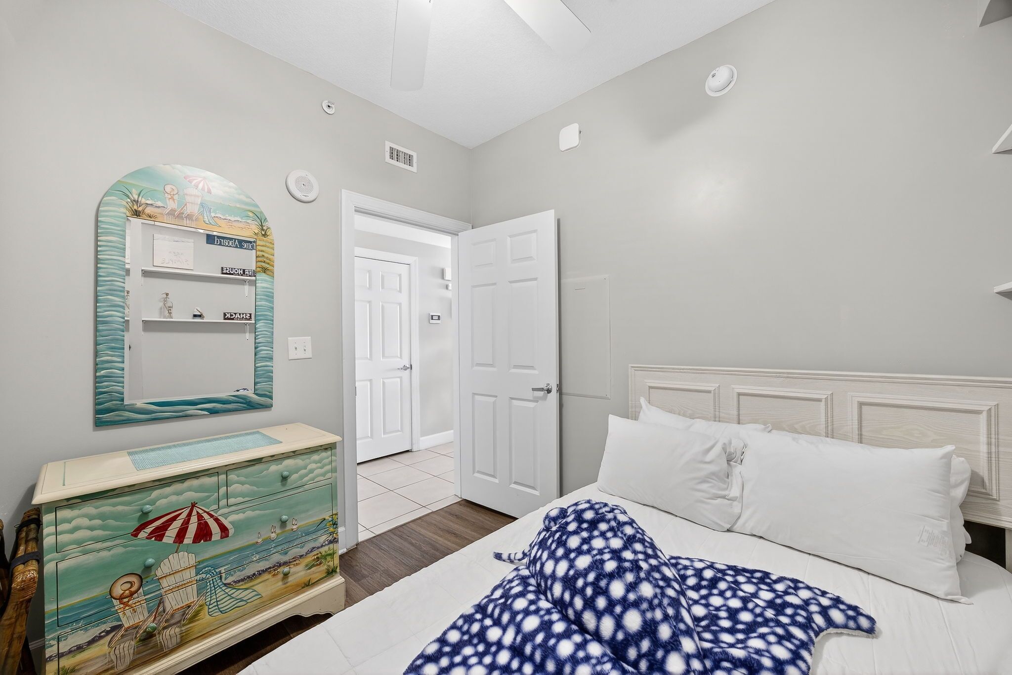 A bedroom with a bed, a dresser with a beach-themed mirror, and a door leading to another room.