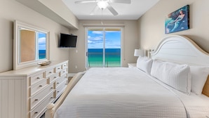 Een slaapkamer met een bed, een dressoir, een plafondventilator, een televisie en een balkon met uitzicht op de oceaan.