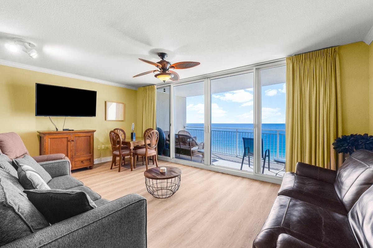 Ein Wohnzimmer mit Sofa, Sessel und Couchtisch. An der Wand ist ein Fernseher angebracht, eine Deckenventilator ist vorhanden, und durch große Fenster hat man Blick aufs Meer.