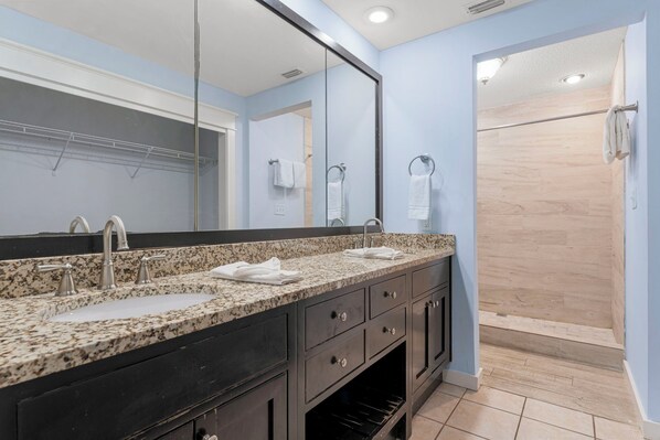 A bathroom with a granite countertop, two sinks, a large mirror, and a shower area. - Seahaven Penthouse (Panama City Beach)