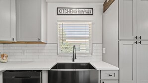 A kitchen with white cabinetry, a stainless steel sink, and a window with blinds. - Sunny Daze Cottage (Panama City Beach)