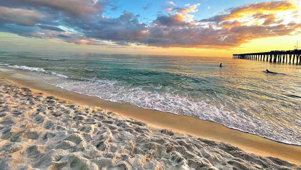 Una platja amb un moll que s'endinsa a l'oceà, on les ones brollen suaument a la riba, i un cel clar per sobre.