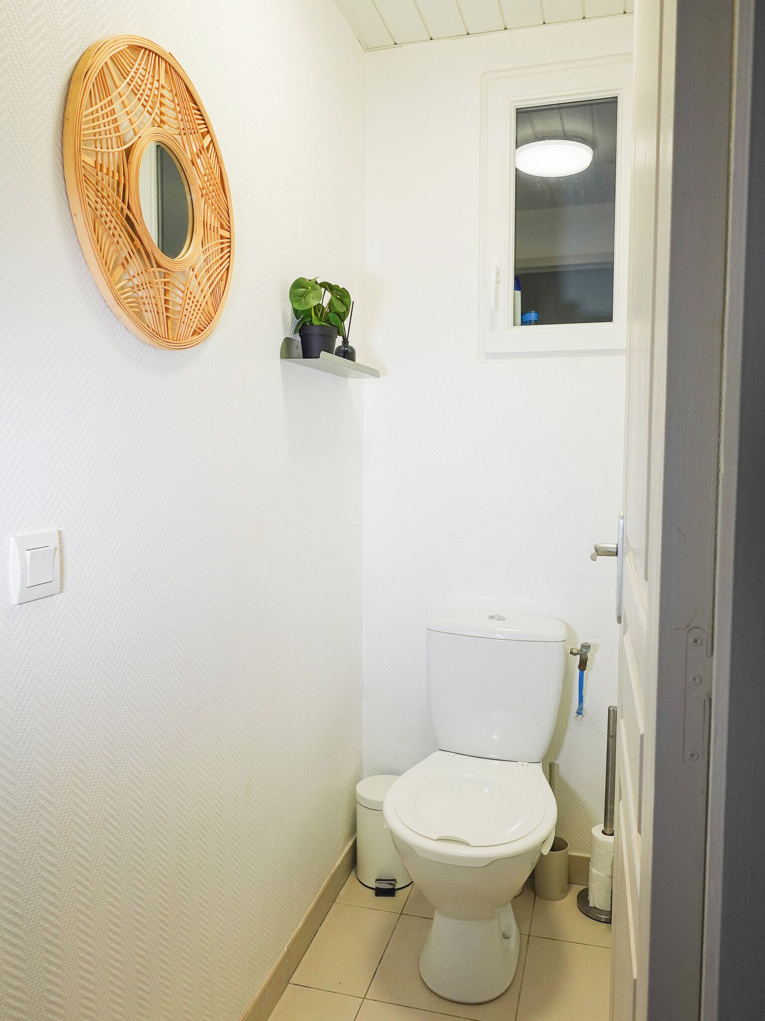 Un baño pequeño y limpio con inodoro blanco, espejo de mimbre, una pequeña estantería con una planta y una ventana sobre el inodoro.