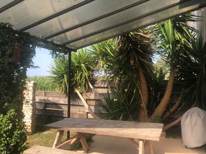 Un espacio al aire libre techado con una mesa de madera, rodeado de abundante vegetación y una valla de madera.
