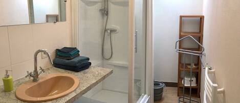 A bathroom with a sink, mirror, shower, and a small wooden shelf.
