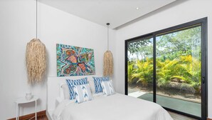 Une chambre moderne avec un grand lit, une petite table de chevet, un tableau abstrait aux couleurs vives et une porte-fenêtre coulissante donnant sur un jardin.