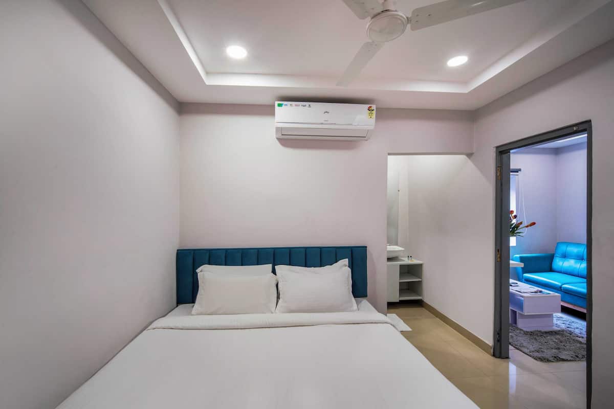 A hotel room with a bed, a ceiling fan, an air conditioner, and a view of a blue sofa in the adjacent room.