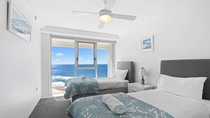 Ett hotellrum med ett stort fönster som ger utsikt över havet, två sängar med blommigt sänglinne, en takfläkt och ett nattduksbord med en lampa.