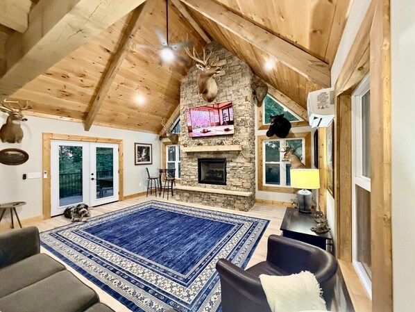 A spacious living room with a stone fireplace, a blue patterned rug, and a ceiling fan.