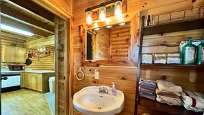 Interior de una cabaña de madera con cocina, un lavabo y un estante con toallas y artículos de aseo.