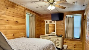 Un dormitorio con paneles de madera, una cama, un aparador, un espejo y un ventilador de techo.