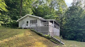 A small cabin with a porch and a staircase leading up to it, surrounded by a grassy hill and dense trees.