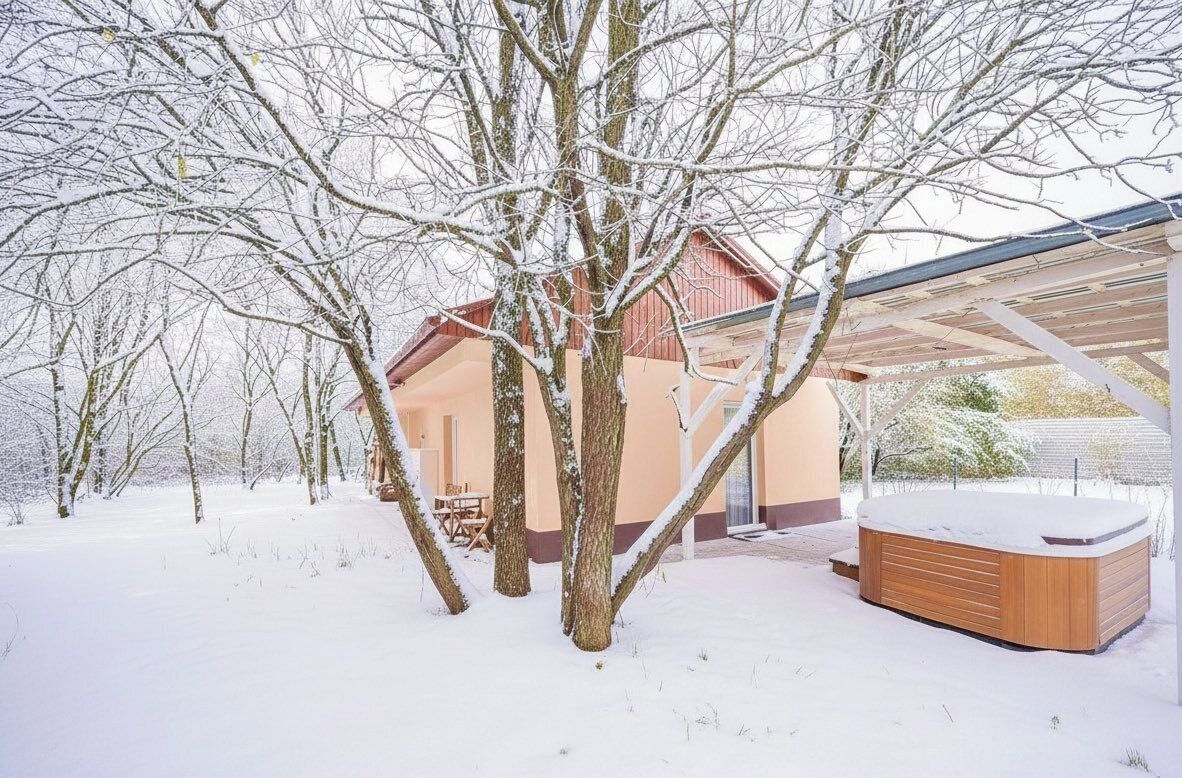 Een besneeuwde buitenruimte met een houten jacuzzi, een gebouw met een schuin dak en kale bomen.