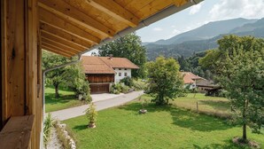 Blick von einem hölzernen Balkon auf eine ländliche Landschaft mit Haus, Bäumen und Bergen.