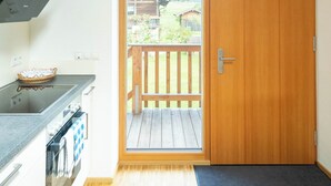 Eine Küche mit integriertem Herd, einer Holztür, die auf einen Balkon führt, und Blick auf ein Haus im Hintergrund.