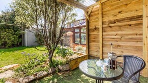 Una terraza de madera con una mesa y sillas de vidrio, rodeada por un jardín con un árbol y una pared de ladrillo.