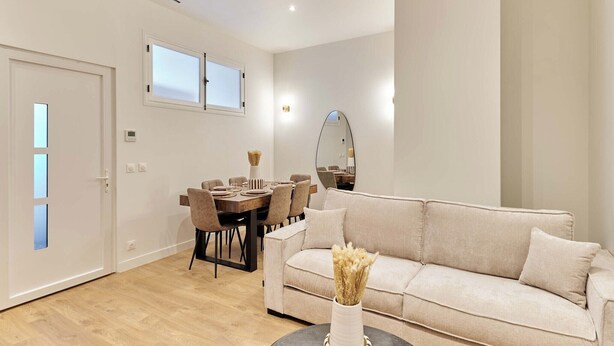 Una sala de estar moderna con un sofá beige, una mesa de comedor con sillas, un espejo y un jarrón con flores secas.