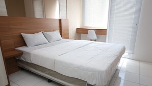 A modern bedroom with a wooden headboard, a white bedspread, and a bedside table with a lamp. - Capital O Iconic Rooms @ Golden City (Bekasi)
