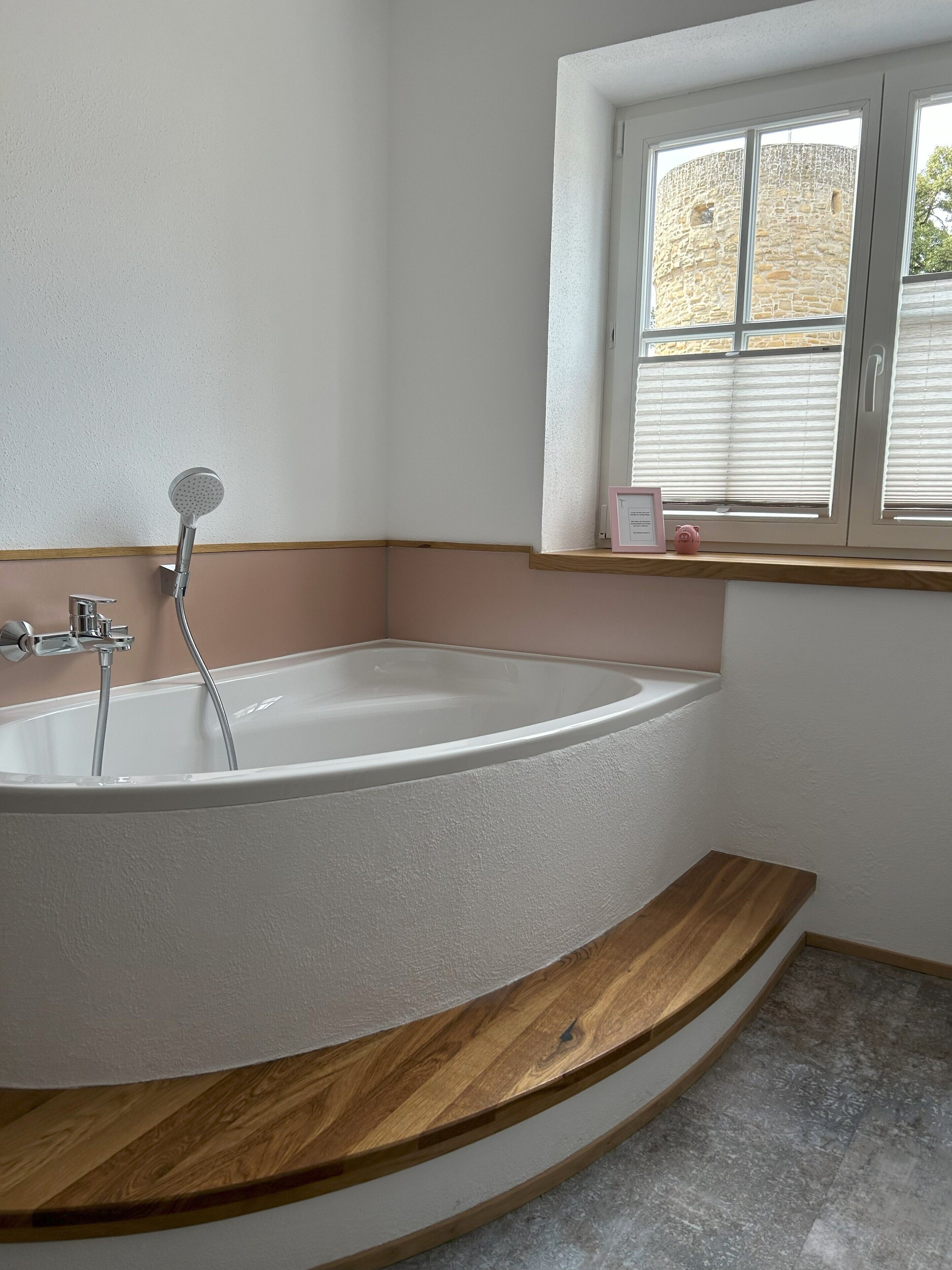 A modern bathroom with a freestanding bathtub, a window with blinds, and a wooden shelf.