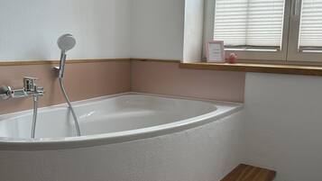 A modern bathroom with a freestanding bathtub, a window with blinds, and a wooden shelf.