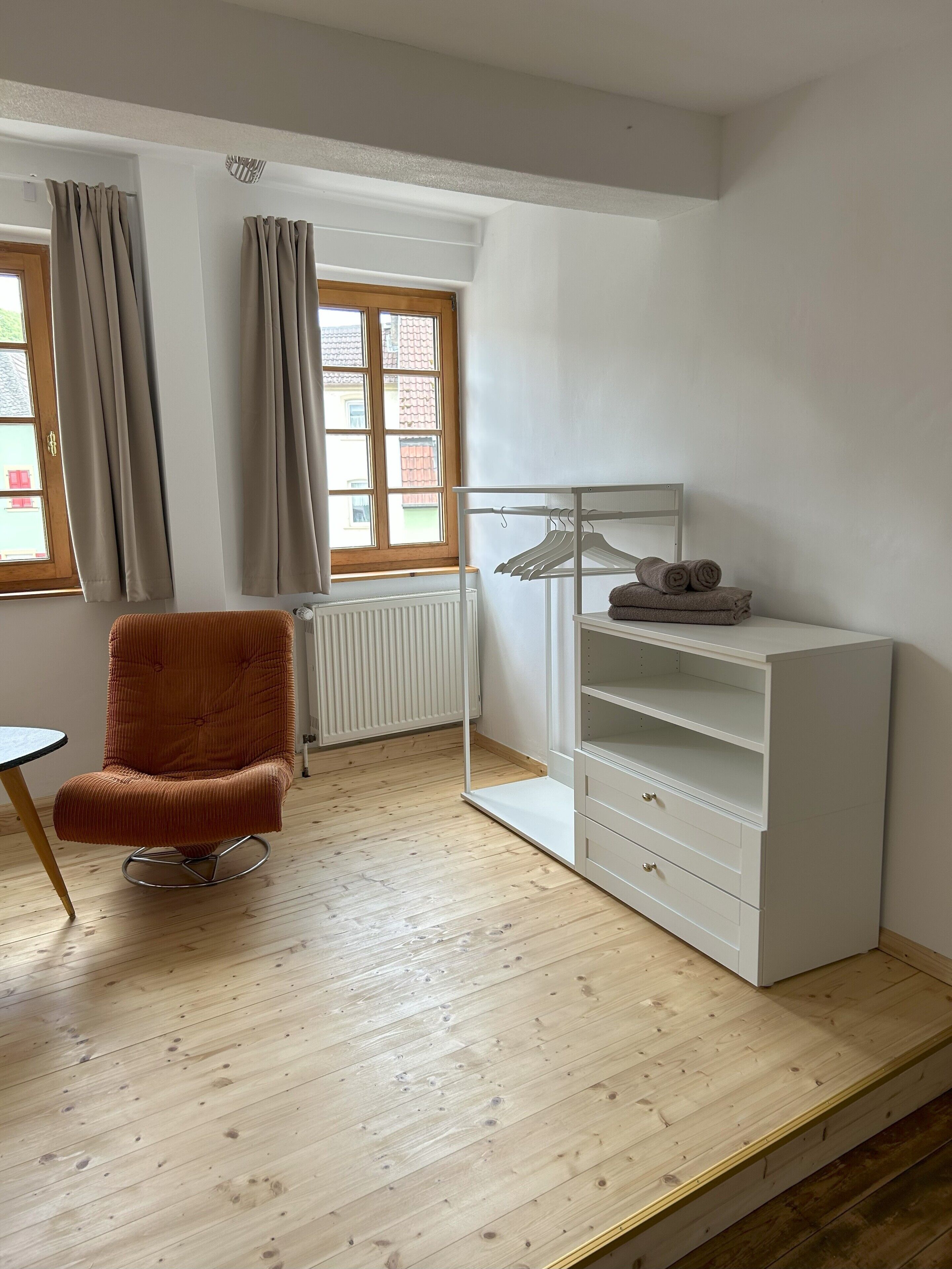 A room with a wooden floor, a brown chair, a white shelving unit, and a window with curtains.