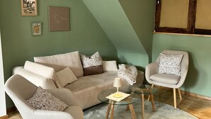 En stue med en beige sofa, to lænestole, et glasbord og en lille lampe. Væggene er dekoreret med billeder, og der er et vindue med træpersienner.