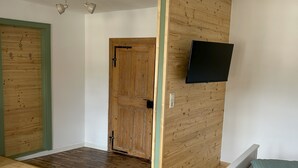 Et værelse med trægulv, en seng med grøn pude, et ophængt tv og en trædør.