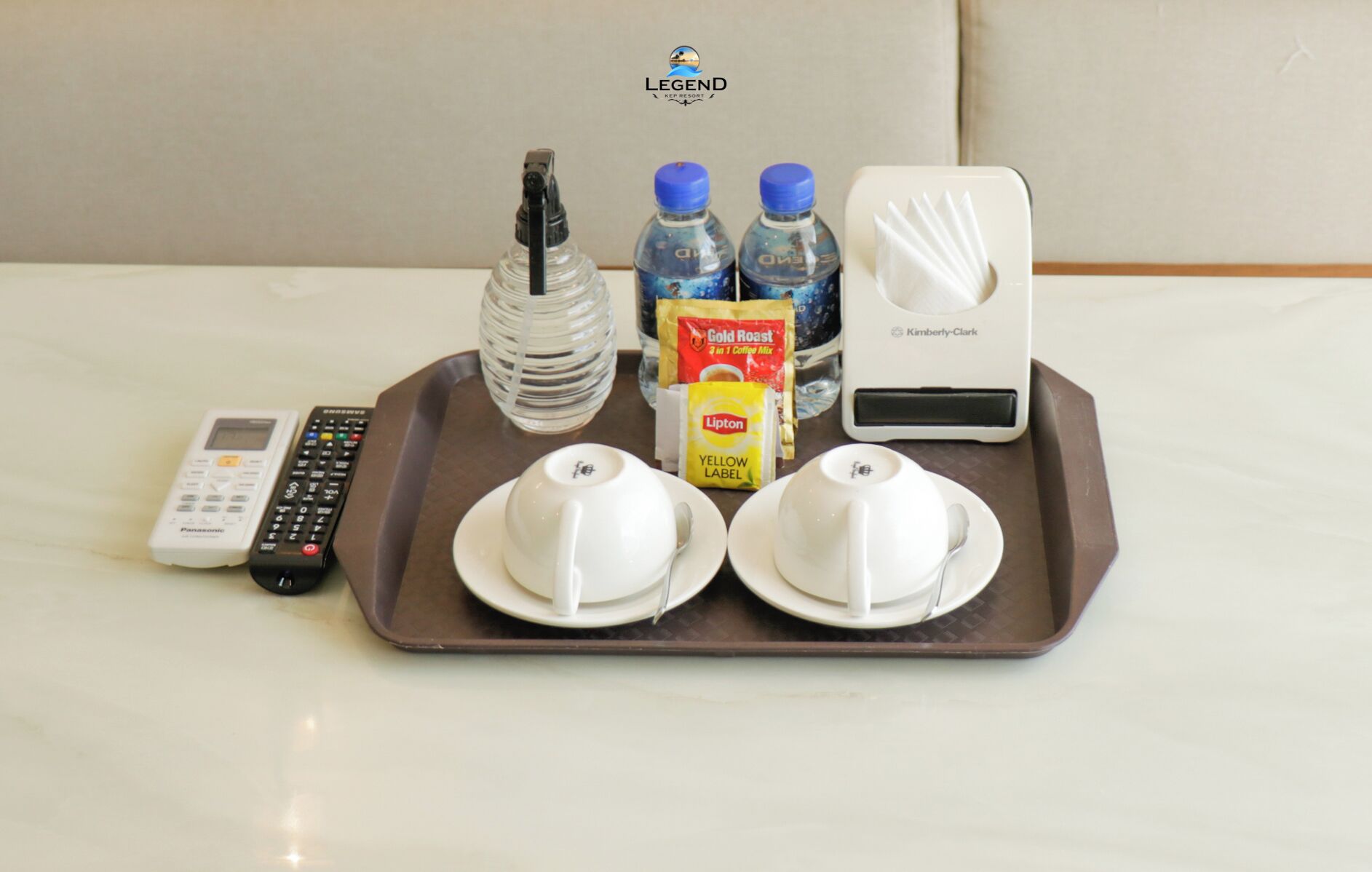 A tray with a remote control, two water bottles, a coffee maker, a Lipton tea packet, and two cups on a table.