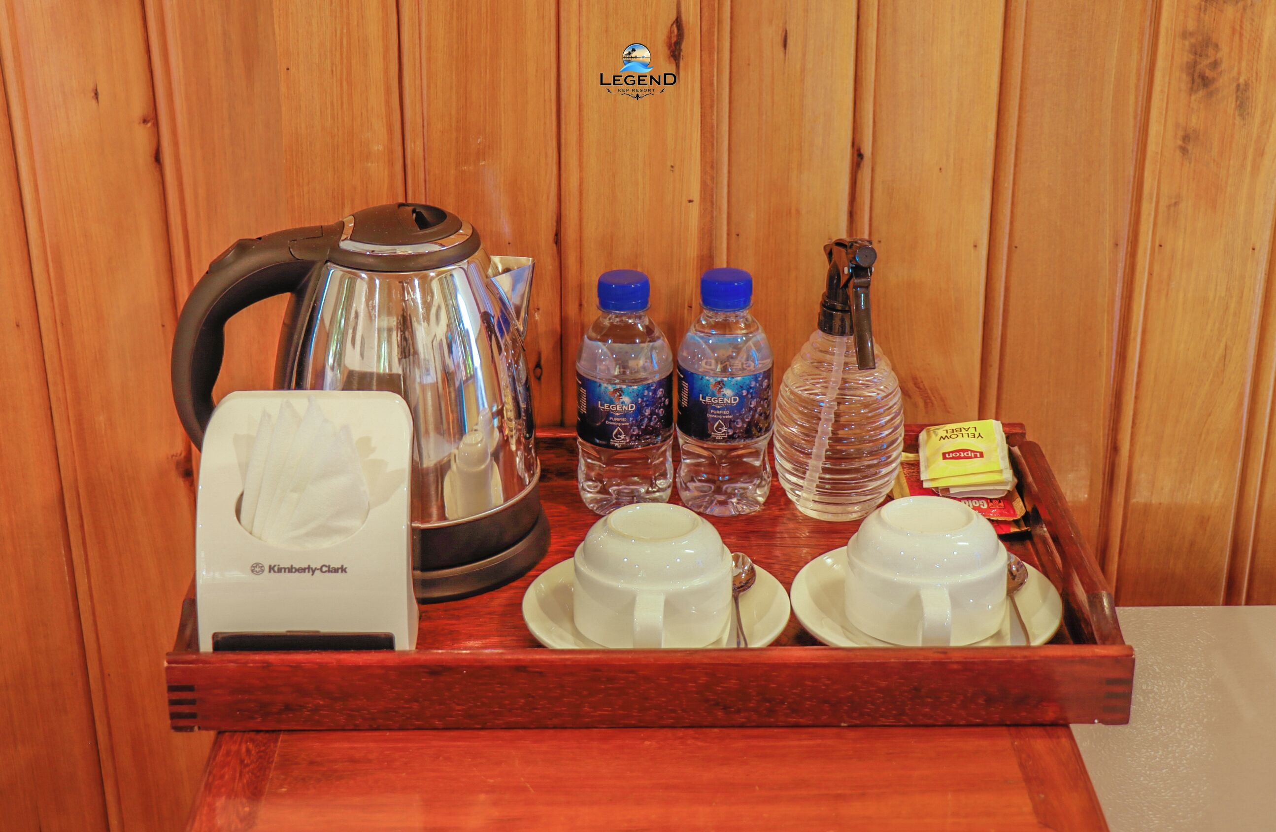 A wooden tray with a tea kettle, two bottles of water, two cups, a tissue box, and a box of tea bags.