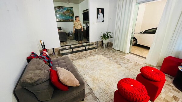 A living room with a grey sofa, red cushions, a white shag rug, and a red ottoman. There is a plant, a mirror reflecting the room, and a white car parked outside.