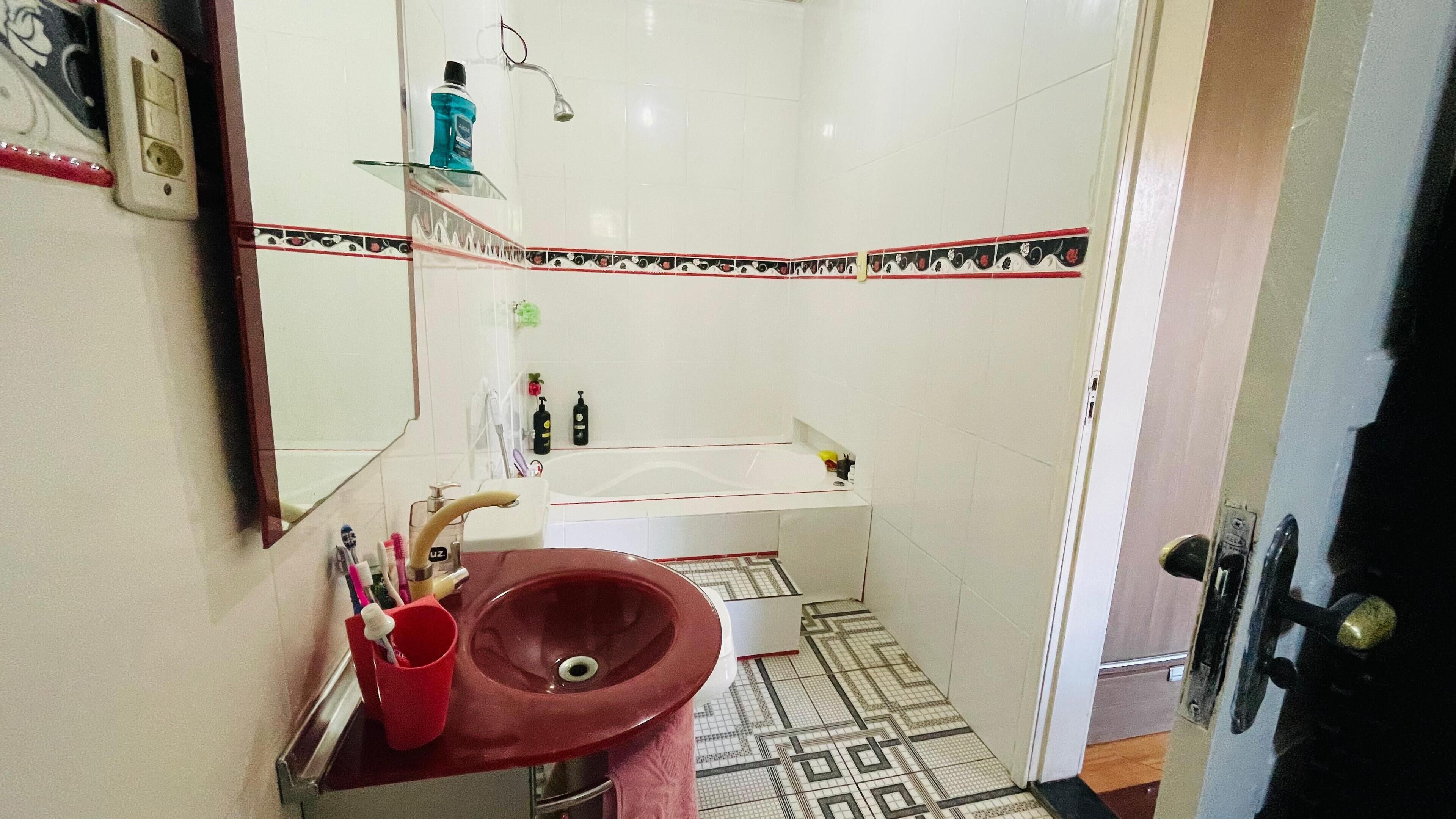 A bathroom with a red sink, a mirror, a white bathtub, and a patterned floor.