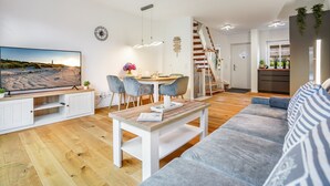 A modern living room with a flat-screen TV, a dining area with a wooden table and chairs, a staircase, and a kitchen area.