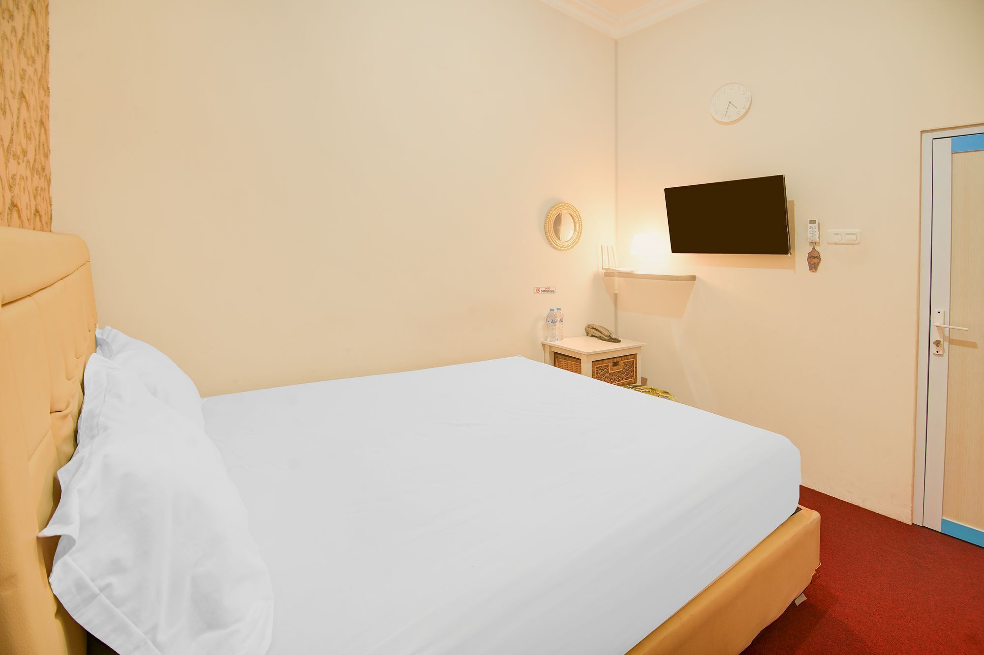 A hotel room with a bed, a bedside table with a lamp, a clock on the wall, and a television mounted above the bed.