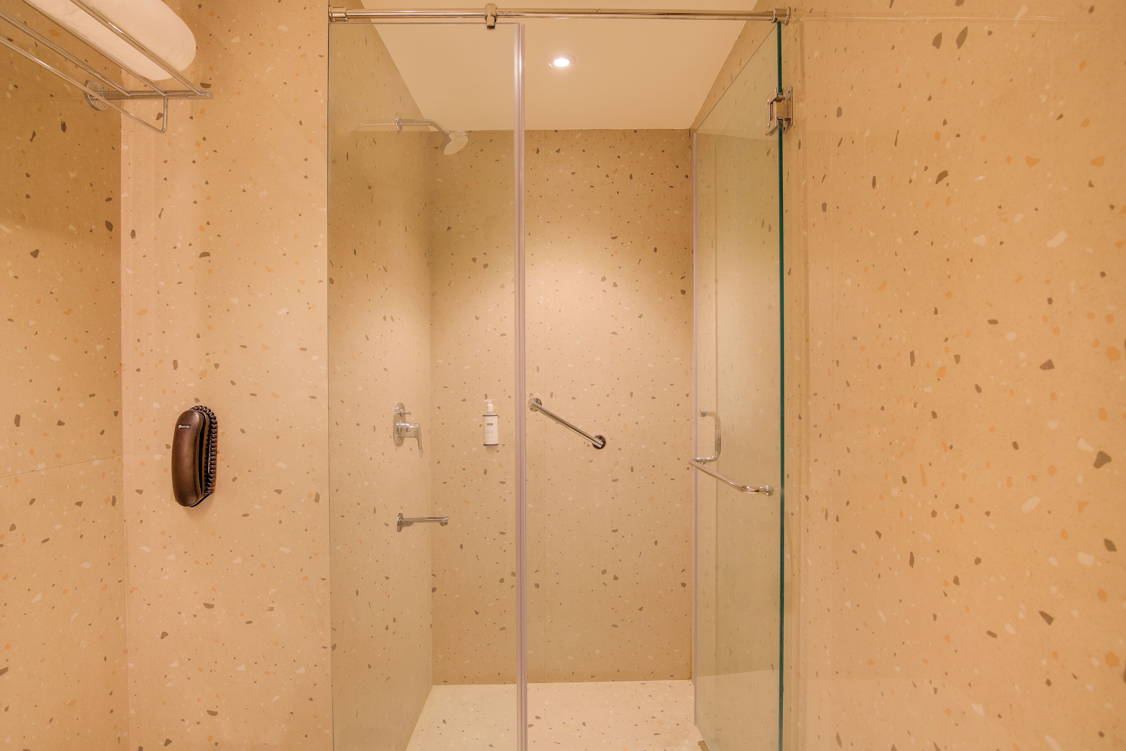 Ducha envuelta en vidrio con pared beige moteada, un perchero y un dispensador de jabón.