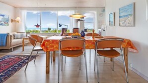 A dining area with a table set for four, surrounded by chairs with orange cushions. There are two pendant lights above the table, and a view of the sea through large windows.