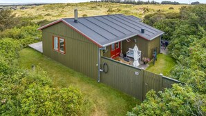 A single-story wooden house with a red door, a covered patio with outdoor furniture, and a fenced yard.