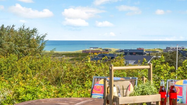 A wooden deck with a table set for dining, overlooking a coastal landscape with houses and the sea.