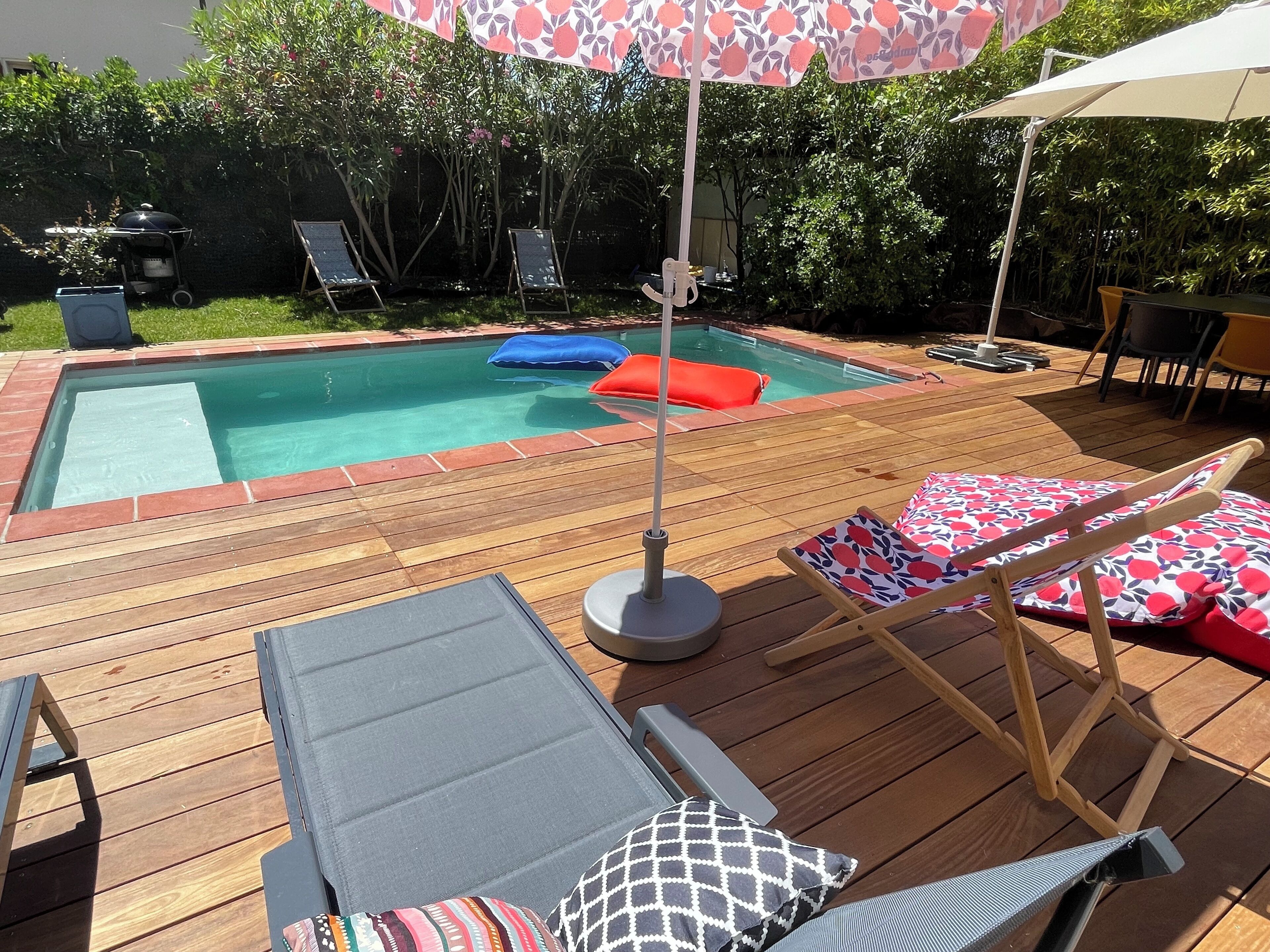 Um deck de madeira com piscina, cadeiras de sol e um guarda-sol.