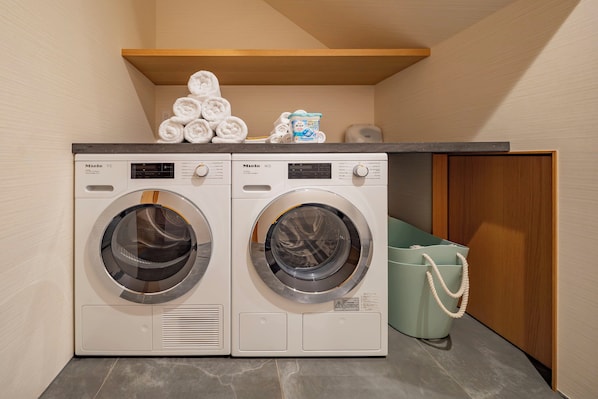 A laundry area with two front-loading washing machines, a stack of neatly folded towels, a container of laundry detergent, and a green laundry basket with a rope handle.
