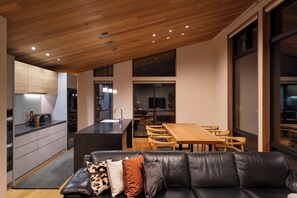 A modern kitchen with a wooden ceiling, a dining area with a wooden table and chairs, and a leather sofa with patterned cushions. - Chalet Opus (Hakuba)