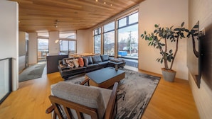 A modern living room with a wooden ceiling, large windows, a black leather sofa, a coffee table, and a potted plant. - Chalet Opus (Hakuba)