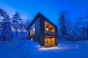 A modern, two-story cabin with a balcony, surrounded by snow-covered trees.