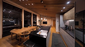 A modern kitchen with a wooden ceiling, large windows, a central island with a sink, and a dining area with wooden chairs. - Chalet Opus (Hakuba)