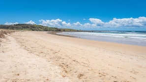 A sandy beach with gentle waves, a clear sky, and a distant shoreline with vegetation.