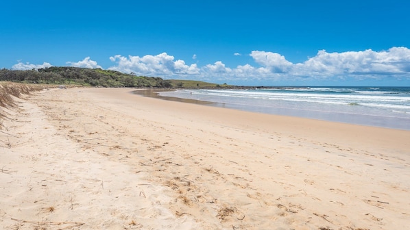 Une plage de sable fin aux vagues douces, un ciel dégagé et une côte lointaine bordée de végétation.