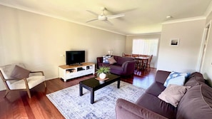 Una sala de estar con televisor de pantalla plana, ventilador de techo, sofá, mesa de centro y un comedor con mesa y sillas.