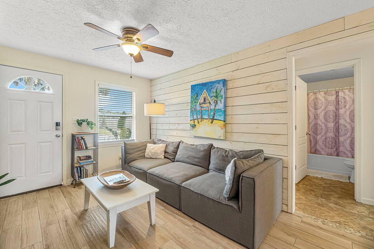 Ein Wohnzimmer mit einem grauen Sofa, einem weißen Couchtisch, einem Deckenventilator, einem Gemälde mit Strandmotiv und einem Bücherregal.