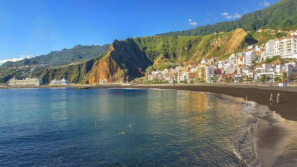 Un pueblo costero con una playa, edificios y montañas al fondo.