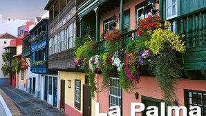 Una calle estrecha flanqueada por edificios de colores, cada uno con balcones decorados con flores colgantes.
